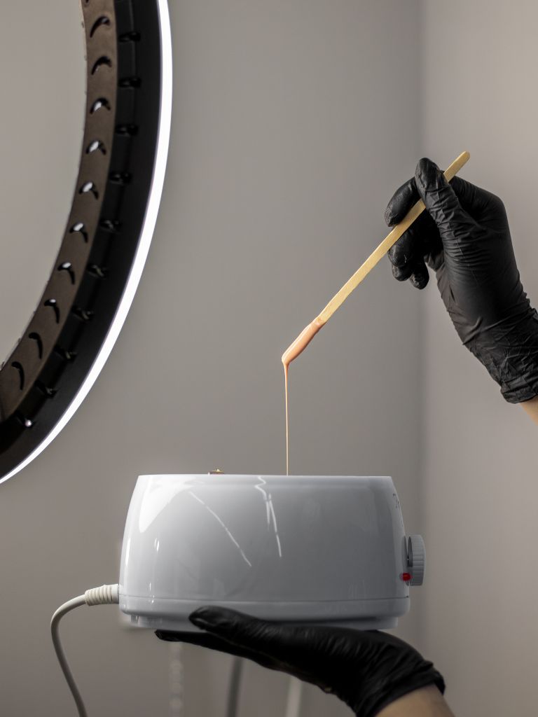 Waxing bei Glamskin: warmes Wachs wird mit einem Holzspatel aus dem Wachswärmer aufgenommen, im Hintergrund ein Ringlicht.